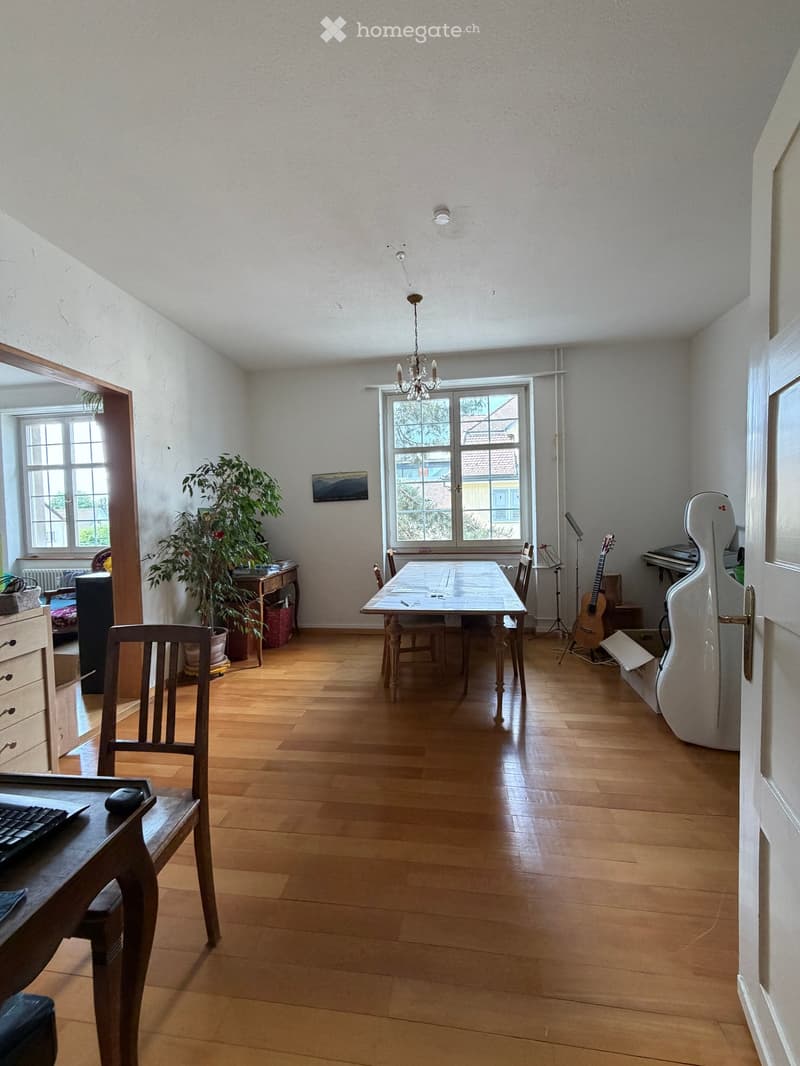 Bright room with wooden flooring, large windows, a dining table with chairs, a guitar, a cello case, a piano, a plant, and a desk with a keyboard and mouse.