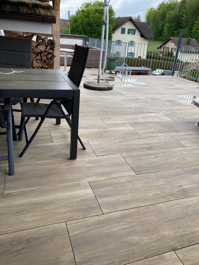 Outdoor patio with a stone floor, a black table and chairs, a stack of firewood, and a view of a house and greenery.