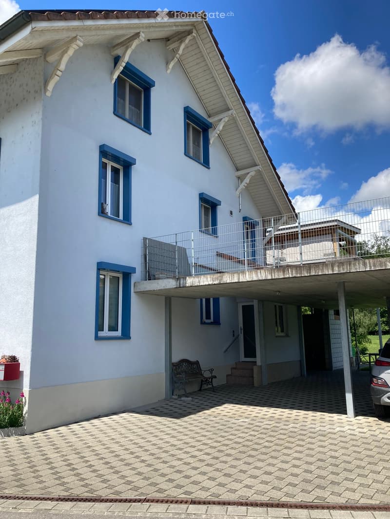 Two-story house with white walls, blue-framed windows, and a balcony on the upper level. Features a tiled driveway, a bench near the entrance, and a small garden area with flowers.
