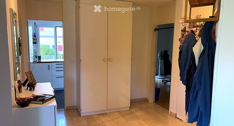 A well-lit hallway with tiled flooring, white walls, and several doors leading to different rooms. A small table with books and decorative items is visible on the left, and a coat rack with jackets is on the right. A fan is seen in one of the adjacent roo