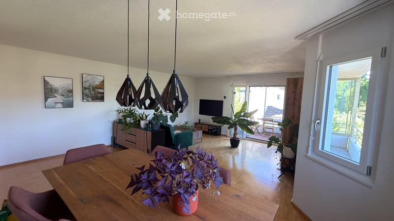 Bright living room with wooden flooring, pendant lights, a large screen, a dining table with chairs, and potted plants. Large windows and a door lead to a balcony area with outdoor furniture.