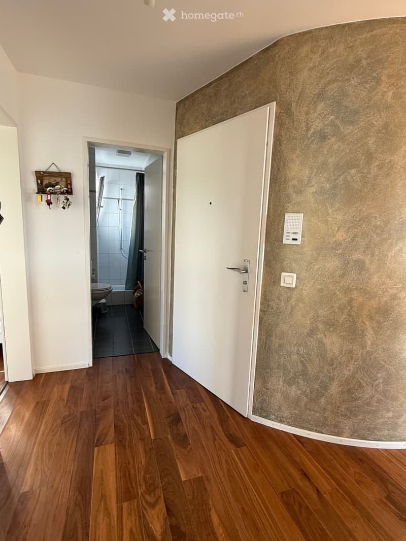 A narrow hallway with wooden flooring, white walls, and a textured wall section. A door leads to a bathroom with white tiles and a shower curtain visible.