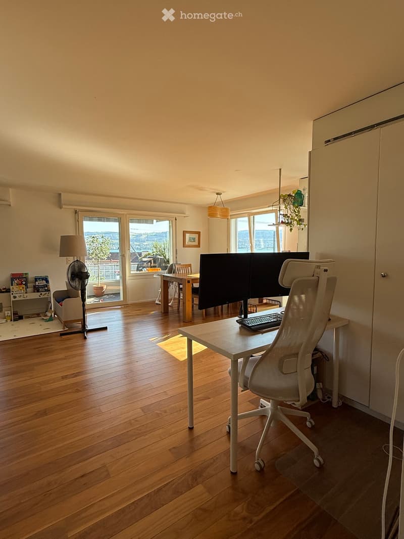 Bright room with wooden flooring, large windows, and a view. Contains a desk with a chair, screen, and keyboard, a small sofa, a fan, and a lamp.