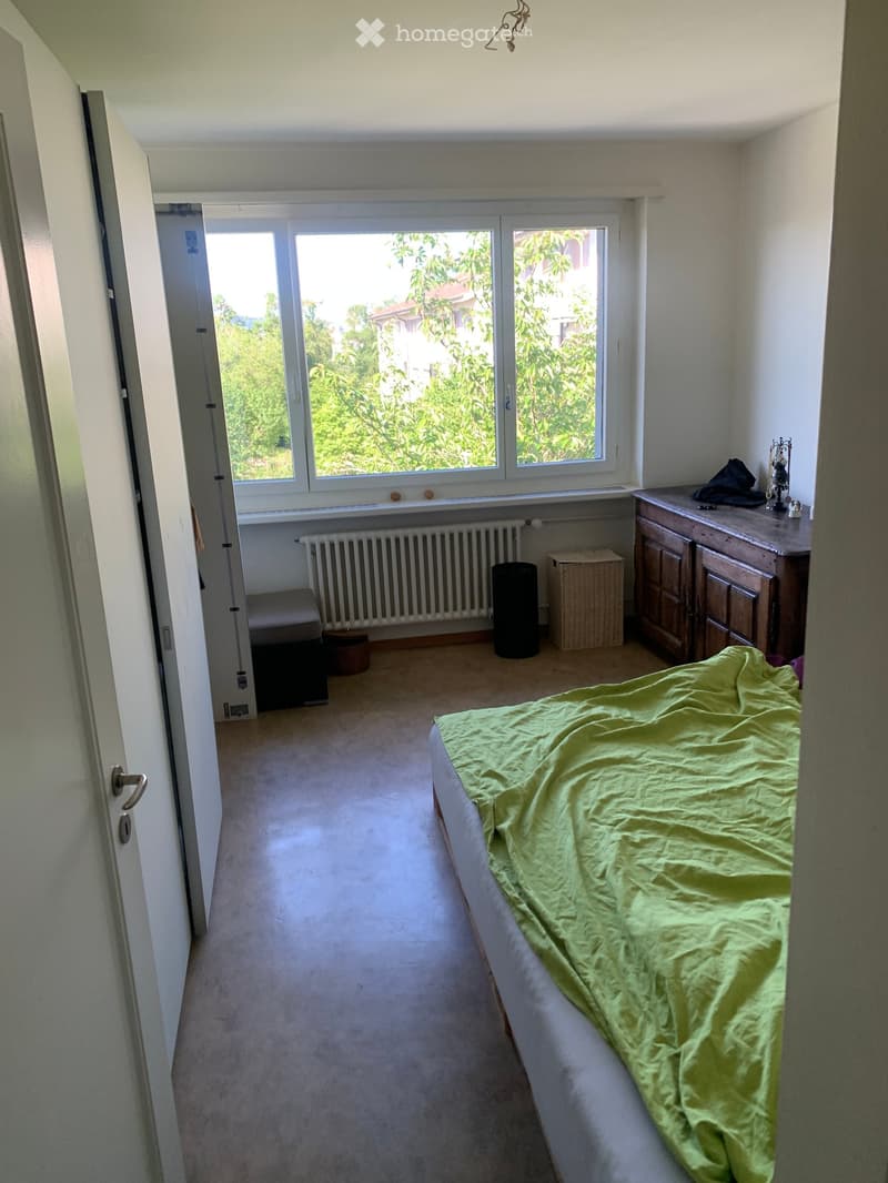 Bright bedroom with a bed covered in a green blanket, a radiator below a large window, a wooden cabinet, a small stool, and a trash bin.