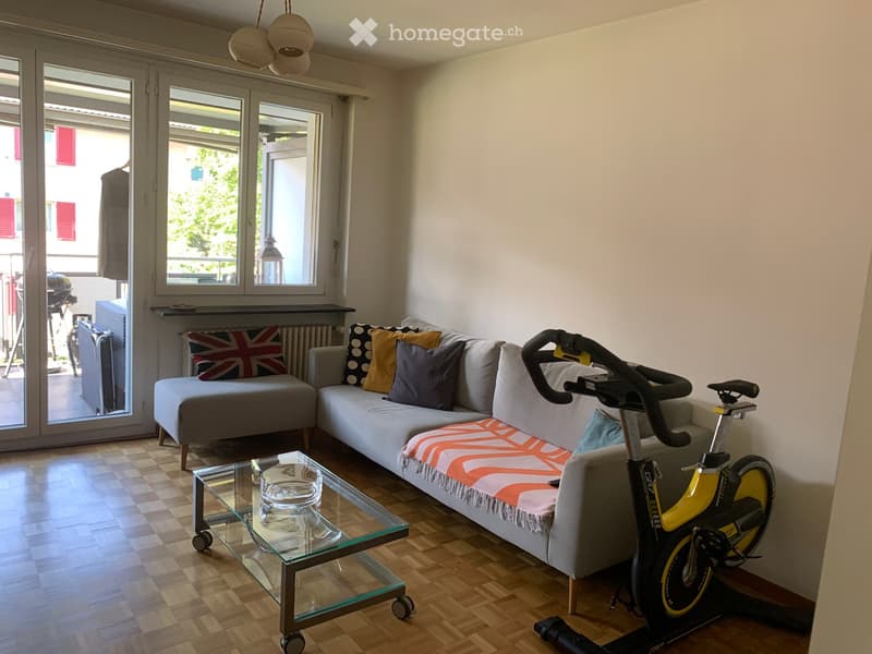 Living room with a grey sectional sofa, glass coffee table, exercise bike, and a small chair. The room has wooden flooring and large windows with a view of the balcony.