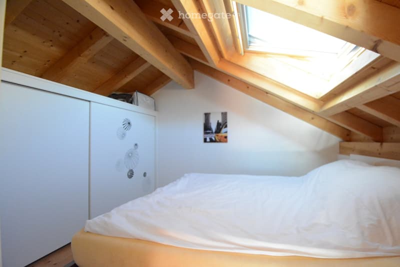 Cozy attic bedroom with wooden slanted ceilings, skylight window, white bed with light-colored bedding, white refrigerator with decorative magnets, and a small picture on the wall.