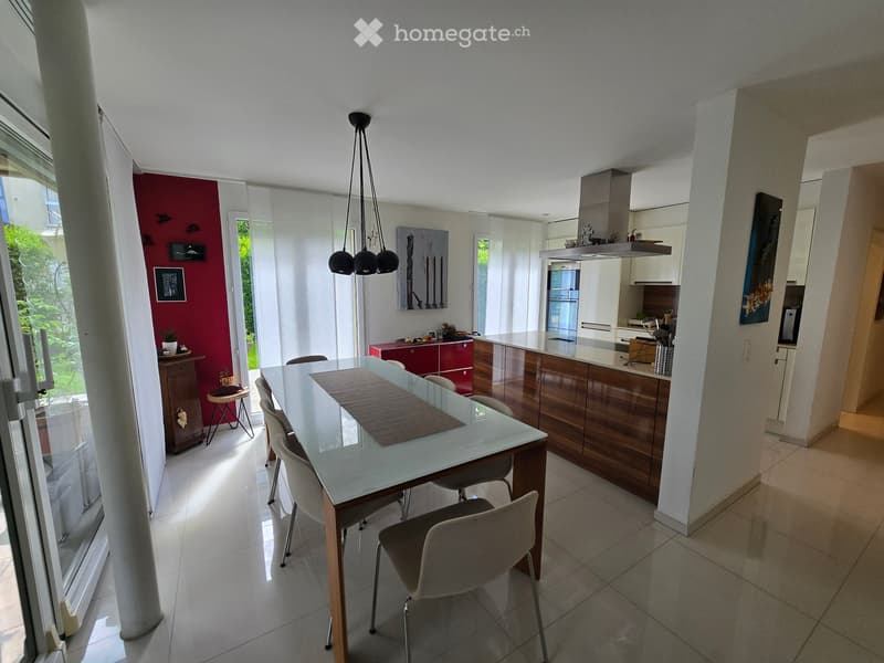Modern open-plan dining and kitchen area with a glass dining table, chairs, kitchen cabinets, and a refrigerator.