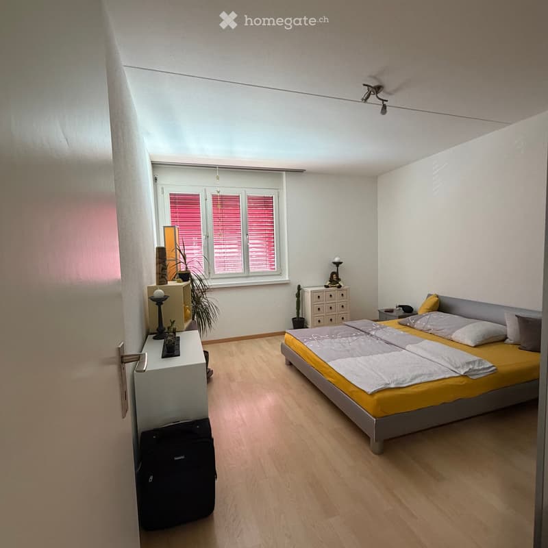 Bright bedroom with wooden flooring, large window with blinds, bed with yellow and grey bedding, nightstand, drawer unit, small table with plants and candle holder, and a suitcase on the floor.