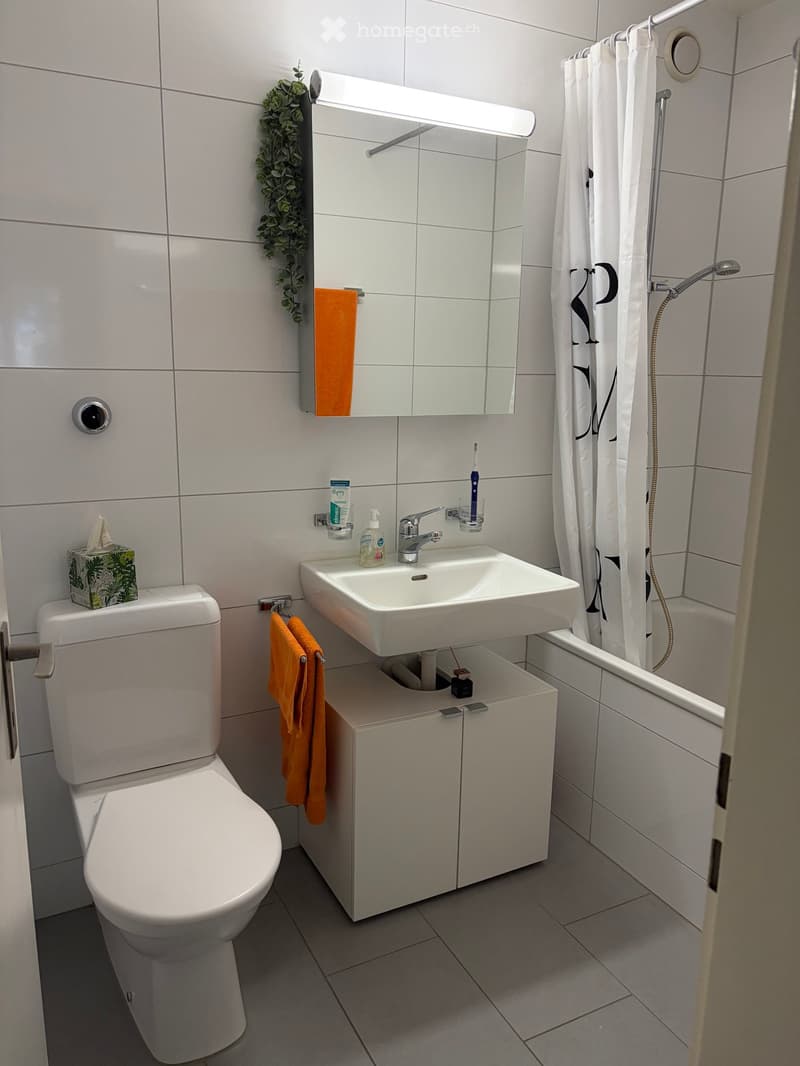Modern bathroom with white tiles, a toilet, a sink with a mirror, a shower area with a curtain, and a small cabinet under the sink.