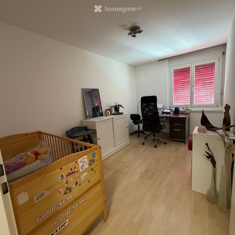 A well-lit room with a crib, desk, office chair, and decorative items, including a plant and a vase.