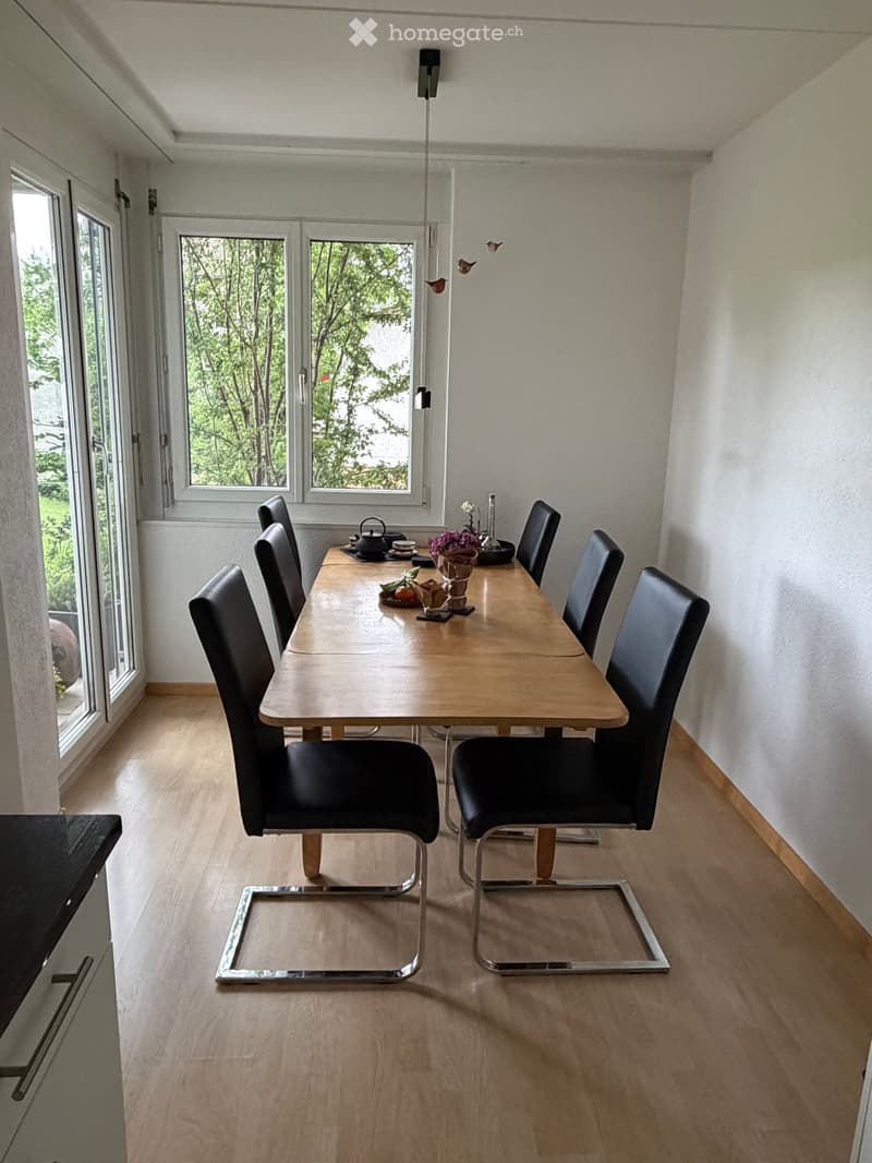 Bright dining area with a wooden table, six black chairs, and decorative items including a teapot, vase with flowers, and candles.