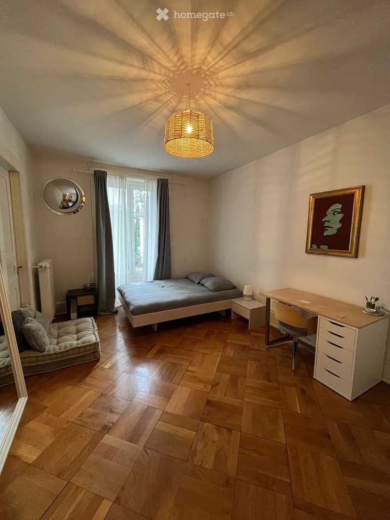 Cozy bedroom with wooden flooring, a bed with grey bedding, a nightstand, a desk with a chair, a dog bed, a round mirror, and a framed picture on the wall.