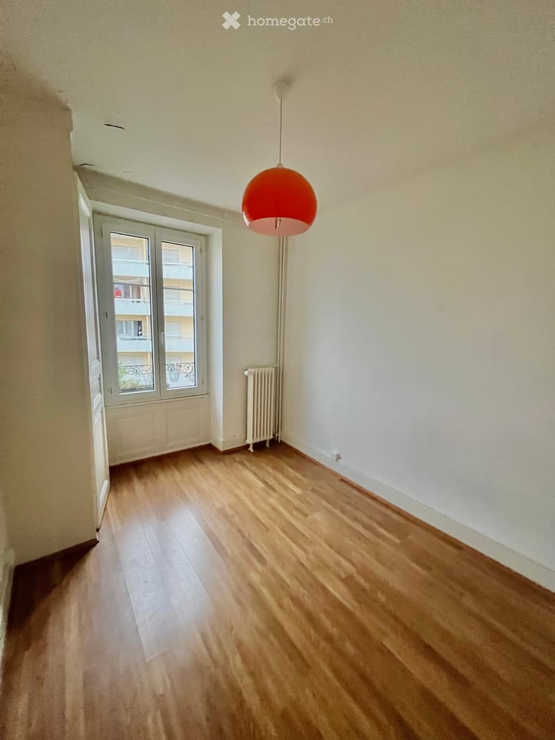 Empty room with wooden flooring, white walls, a radiator, and a hanging orange lamp. A window with glass panels opens to a view of a building exterior.