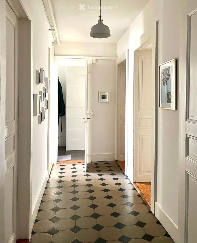 A long, narrow hallway with a geometric tile floor, white walls, and several doors leading to different rooms. A pendant light hangs from the ceiling, and framed pictures are displayed on the walls.