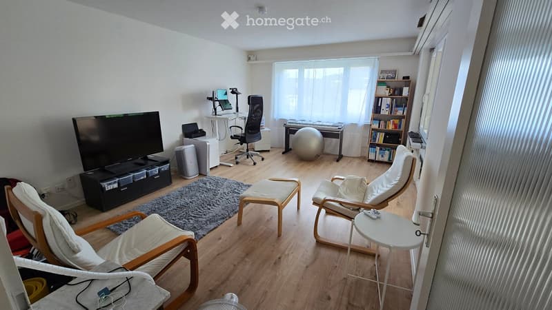 A modern living room with a large screen, comfortable chairs, a small table, a laptop on a desk, a bookshelf, and a piano. The room features wooden flooring and large windows with sheer curtains.