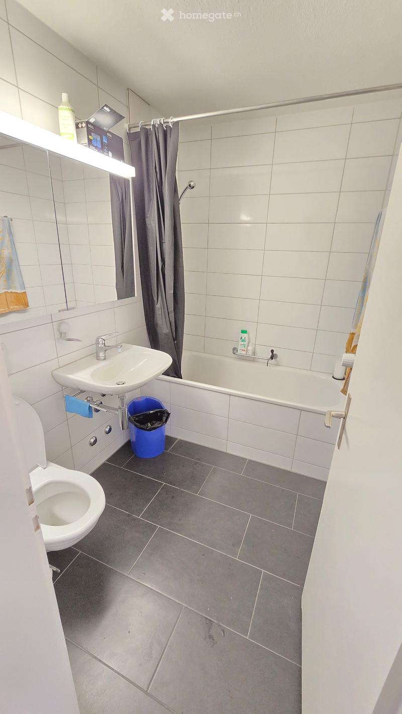 Modern bathroom with white tiles, a toilet, sink, bathtub, and a shower curtain. Includes a mirror, towel, and a trash bin.