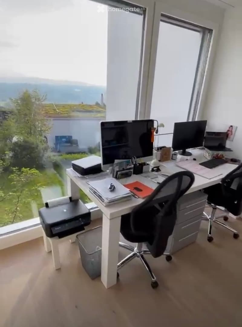 Modern office space with a large window offering a scenic view, equipped with a desk, two chairs, a printer, and multiple computer screens.