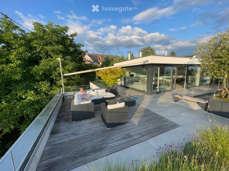 Modern outdoor terrace with wooden decking, wicker seating, glass-enclosed living space, and lush greenery.
