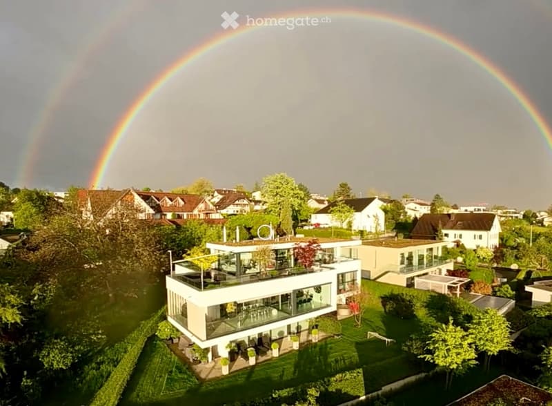 Modern two-story house with large windows and a flat roof, surrounded by greenery and situated near a body of water with rainbows in the sky.