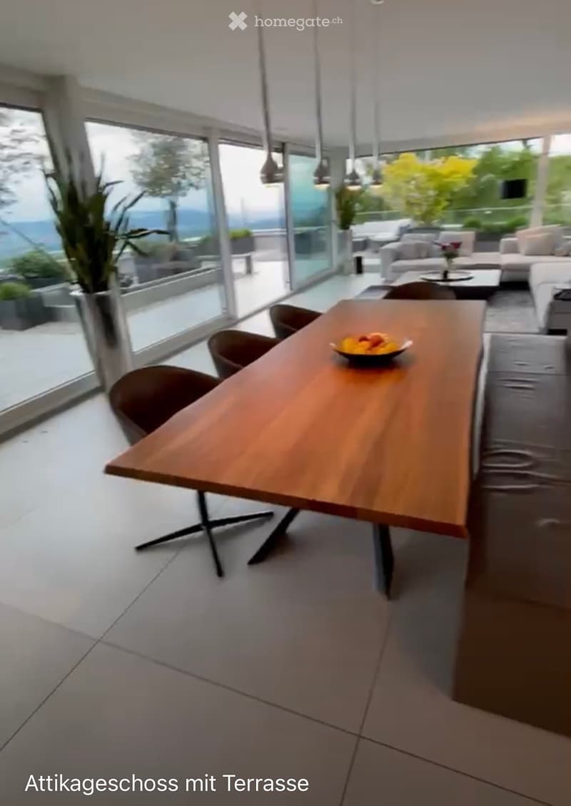 Modern open-plan living and dining area with large windows offering a scenic view, featuring a wooden dining table, chairs, a sofa, and a coffee table.