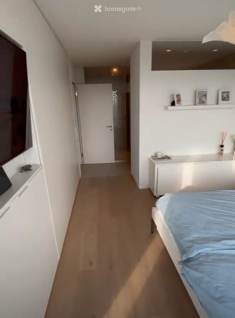 Modern bedroom with a white bed, blue bedding, nightstand, and shelves with picture frames. The room has light-colored walls and wooden flooring.