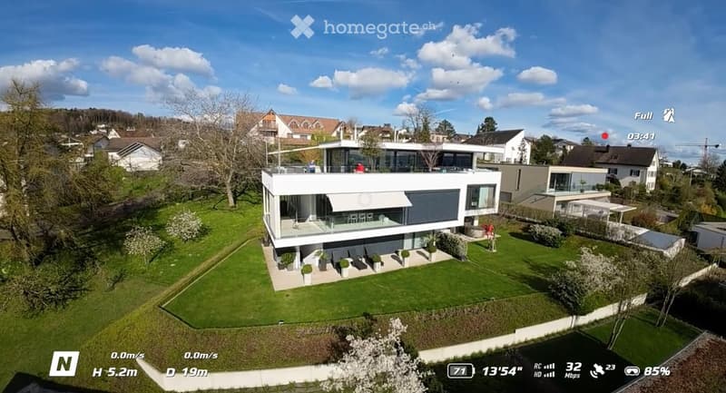 Modern two-story house with a flat roof, large windows, and a spacious lawn. The house features a raised ground floor and is surrounded by greenery and neighboring houses.