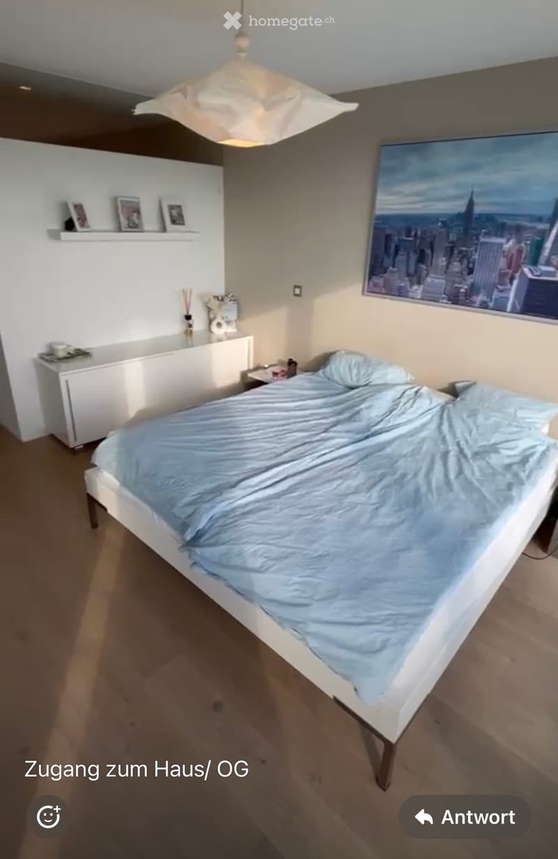 Modern bedroom with a white bed, light blue bedding, a nightstand, and a shelf with picture frames and decorative items.