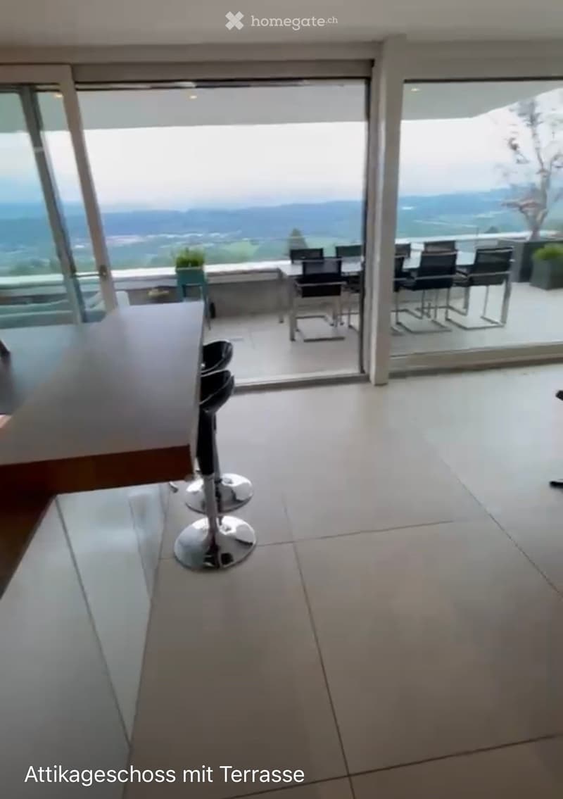 Modern interior with large glass doors leading to a terrace, offering a scenic view. Includes a stool and chairs on the terrace.
