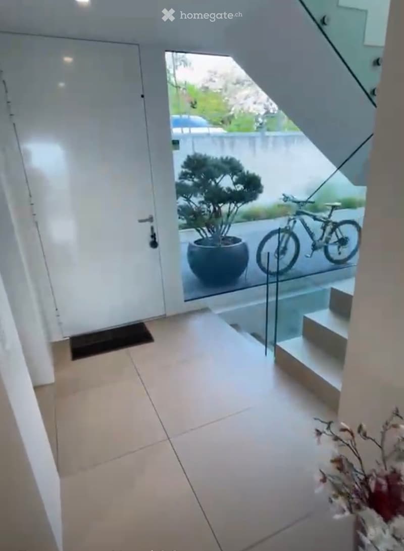 Modern hallway with light-colored tiles, white walls, and a glass door leading to an outdoor area with a potted plant and a bicycle.