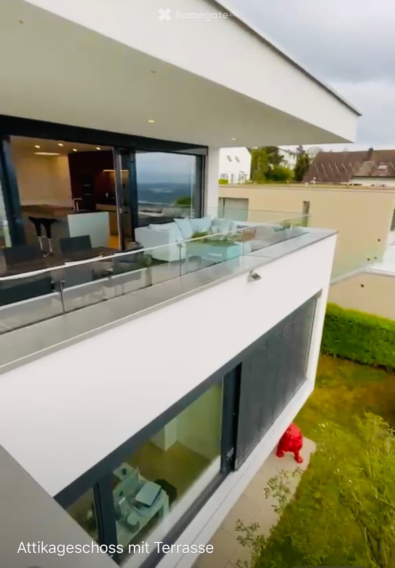 Modern rooftop terrace with glass railing, overlooking a green lawn and neighboring buildings. The terrace is part of an attic floor with large windows and a sleek design.