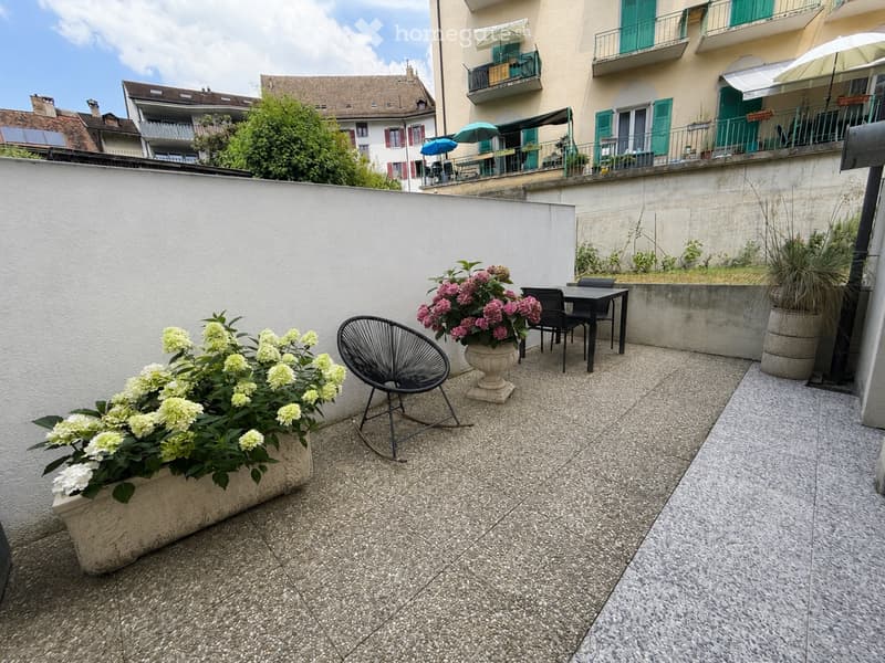 Balcony with a tiled floor, two flower pots with pink and white flowers, a black modern chair, and a small black table with two black chairs.