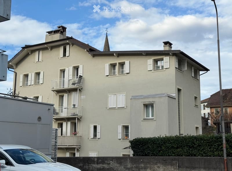 A three-story residential building with light beige walls, multiple windows with white shutters, and balconies on the upper floors.