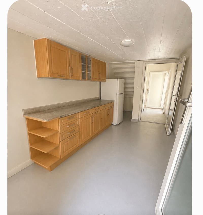 Empty kitchen with light wooden cabinets, granite countertop, and a white refrigerator. The space is clean and well-lit with a visible doorway leading to another room.