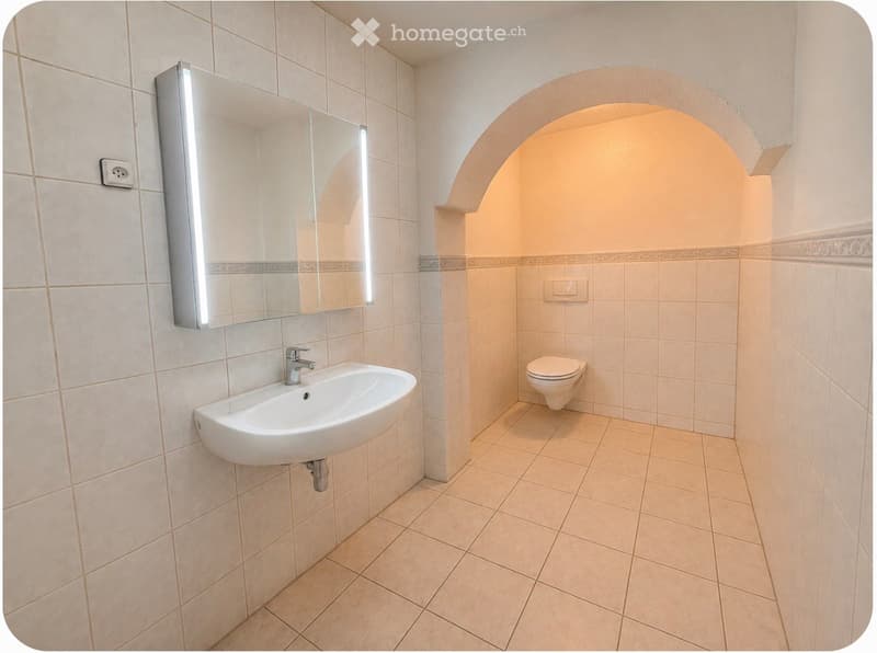 Modern bathroom with beige tiles, a white sink, a toilet, and a large mirror with built-in lighting.