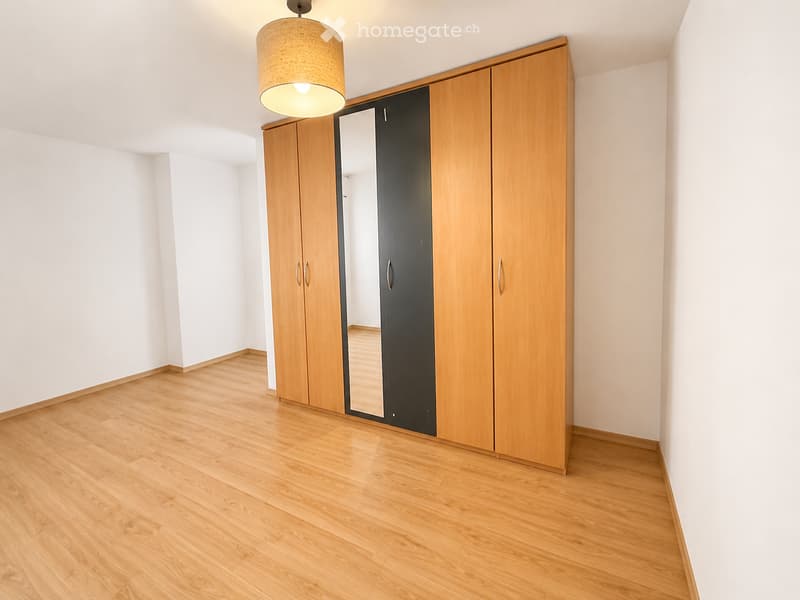 Empty room with light wooden flooring, white walls, and a large wooden wardrobe with mirrored doors.