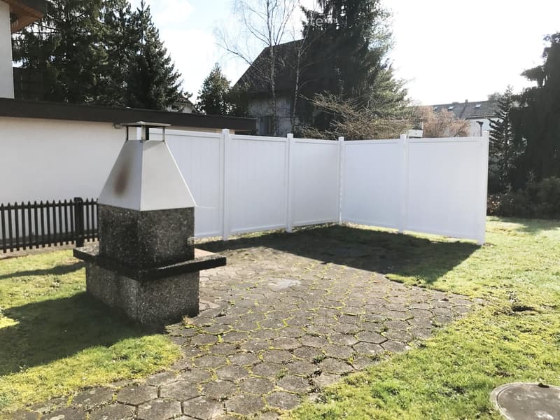 A small outdoor area with a stone chimney, paved ground, white fence, and surrounding greenery.