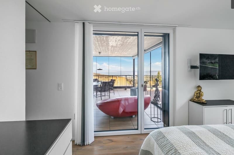 Modern bedroom with wooden flooring, large glass doors leading to a balcony with a scenic view, a bed, a red sofa, a dining table with chairs, and decorative statues.