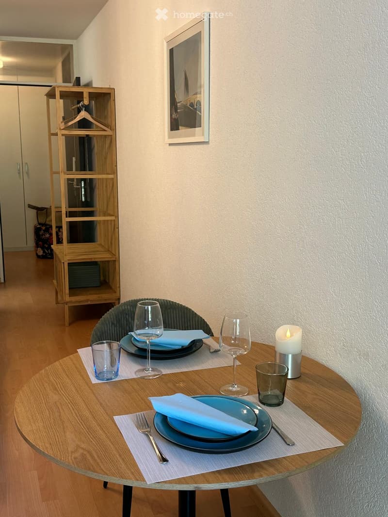 A small, neatly arranged dining area with a round wooden table set for two, featuring plates, wine glasses, cutlery, and a lit candle. A wooden shelving unit is visible in the background.