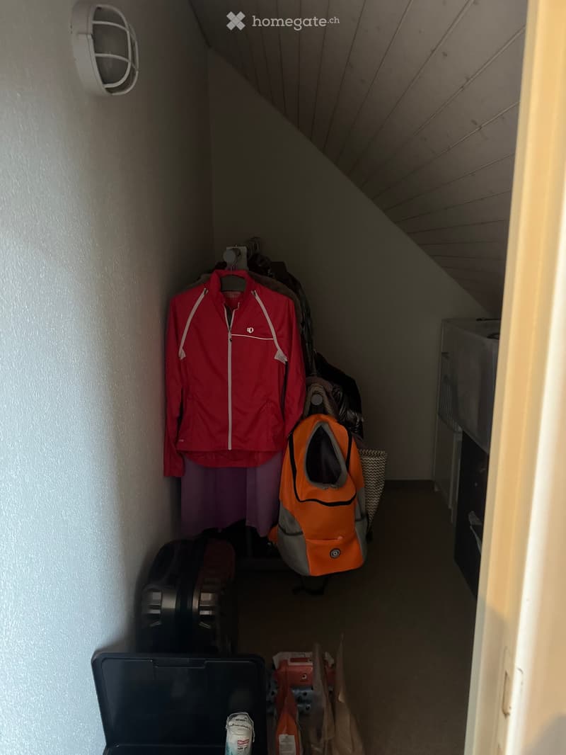 Narrow hallway with slanted ceiling, clothes hanging on a rack, and various items including a backpack, shoes, and a small shelf with items.