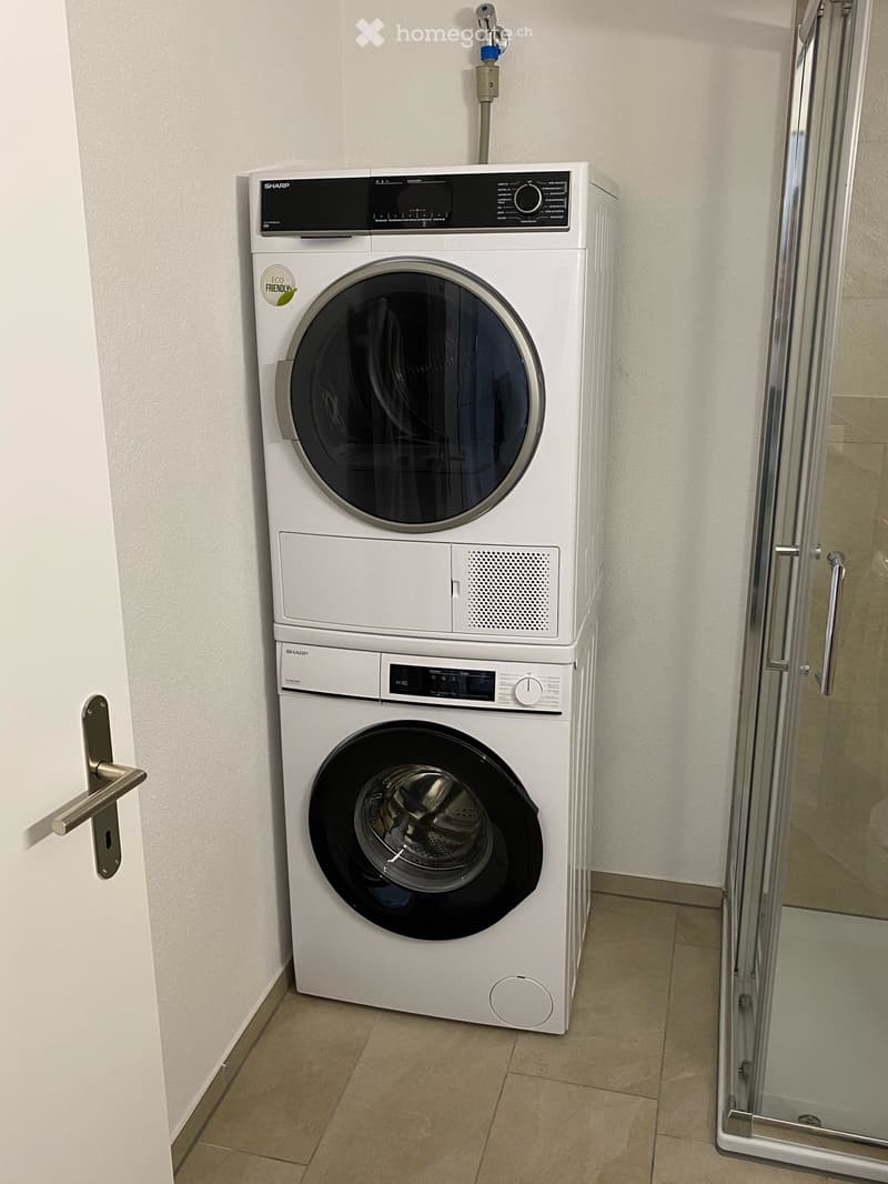 Bathroom with a white washing machine and dryer unit, glass shower enclosure, tiled floor, and white walls.
