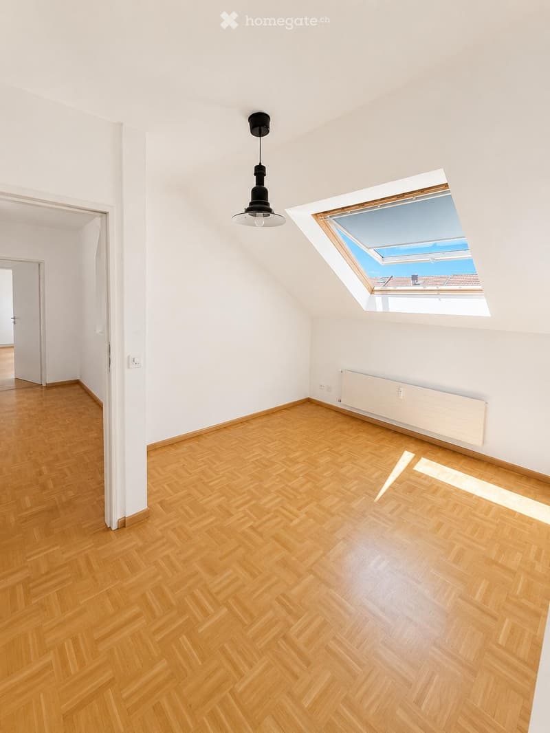 Empty room with wooden herringbone flooring, white walls, a skylight window, and a single hanging light fixture.