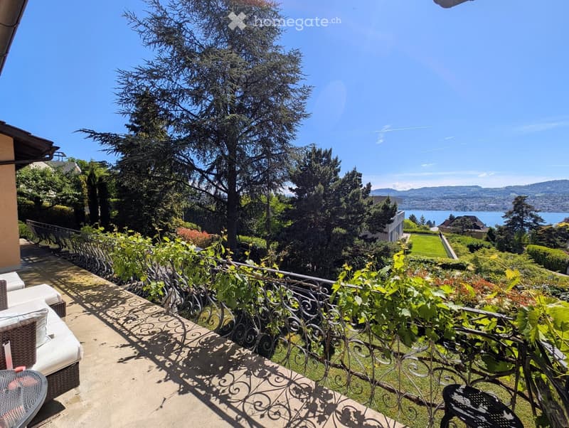 Balcony with a view of a lake and surrounding greenery, featuring a white sofa, a table, and a decorative railing.
