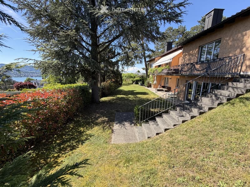 A two-story house with a balcony, surrounded by a well-maintained garden with grass, shrubs, and trees. The house features large windows and a staircase leading to the balcony.