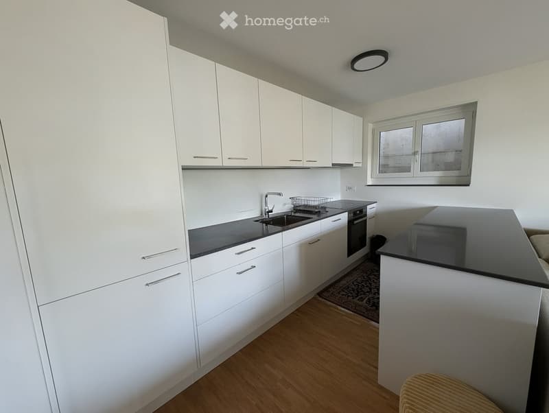 Modern kitchen with white cabinets, black countertops, a sink, and an oven. The space features wooden flooring and a small window above the sink.