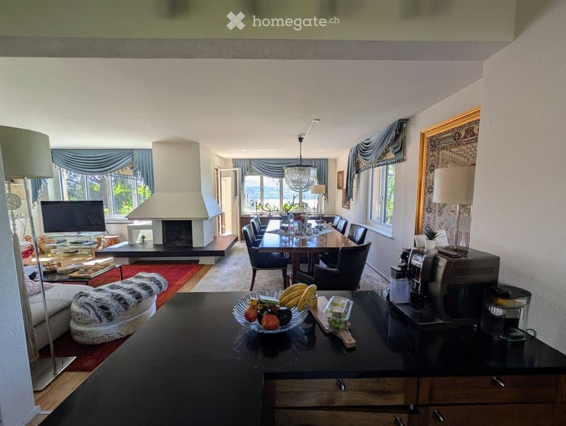 Open-plan living and dining area with a fireplace, large windows offering a scenic view, a sofa, coffee table, dining table with chairs, and a coffee machine on the counter.