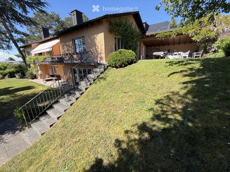 Two-story house with a raised ground floor, surrounded by a well-maintained lawn and steps leading up to a balcony area.