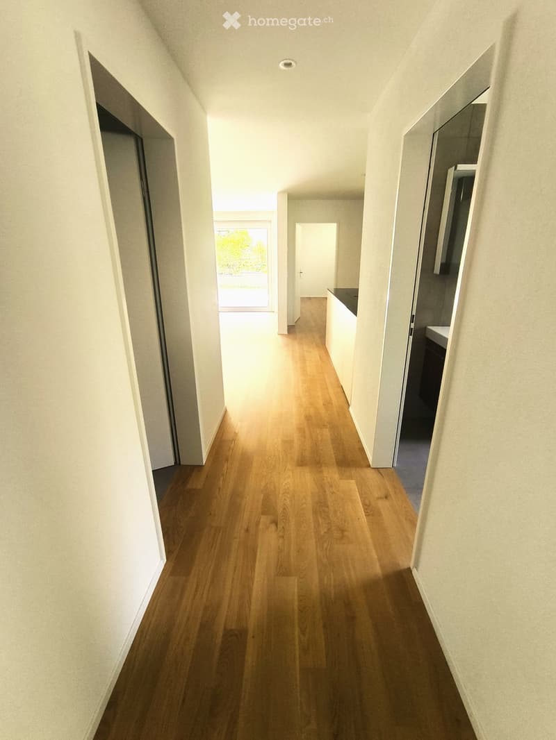 A long, narrow hallway with light wooden flooring and white walls, leading to an open area with natural light and a view of greenery outside.