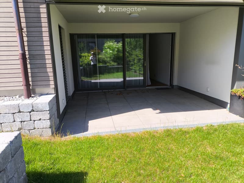 Modern outdoor patio area with tiled flooring, glass sliding doors leading to a house, stone retaining wall, and well-maintained lawn.