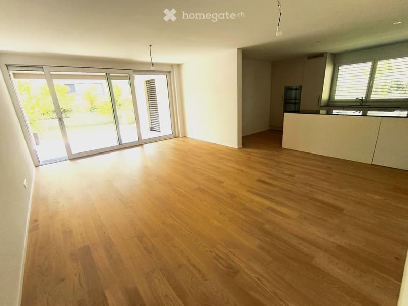 Empty room with wooden flooring, large sliding glass doors leading to a garden, and a small kitchen area visible in the background.