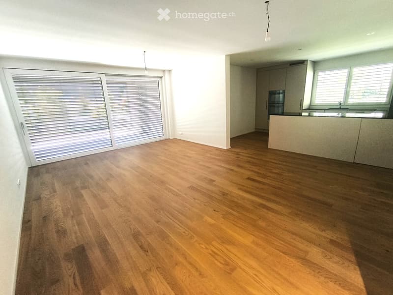 Empty room with wooden flooring, white walls, and large windows with blinds. The adjacent kitchen area has cabinets and a countertop.